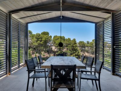 Vaste terrasse couverte avec table à manger en bois rustique et chaises sombres, encadrée de panneaux à persiennes offrant intimité et vue dégagée sur la végétation méditerranéenne.