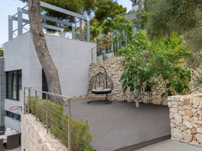 Vaste terrasse extérieure en composite foncé, agrémentée d'un fauteuil suspendu et encadrée de murs en pierre naturelle. La végétation méditerranéenne et l'architecture moderne se complètent harmonieusement.