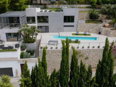 Vue aérienne de la villa moderne avec piscine rectangulaire et terrasses étagées. La façade grise, les jardins en restanques de pierre et les vastes espaces extérieurs sont visibles.