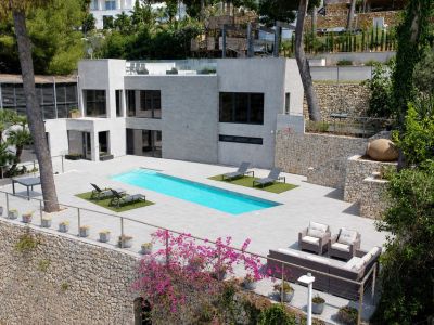 Vue aérienne de la villa moderne, avec sa façade grise épurée, sa piscine privée rectangulaire et ses vastes terrasses carrelées. Murs en pierre et végétation méditerranéenne luxuriante.