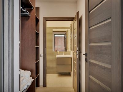 Vue d'un couloir vers une salle de bain moderne avec meuble-lavabo, miroir et murs carrelés. Une armoire ouverte en bois foncé avec étagères est visible à gauche.