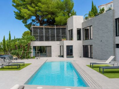 Vue extérieure de la villa avec piscine privée rectangulaire, terrasses carrelées grises et zones de gazon artificiel. L'architecture moderne et une terrasse supérieure à lames se distinguent.