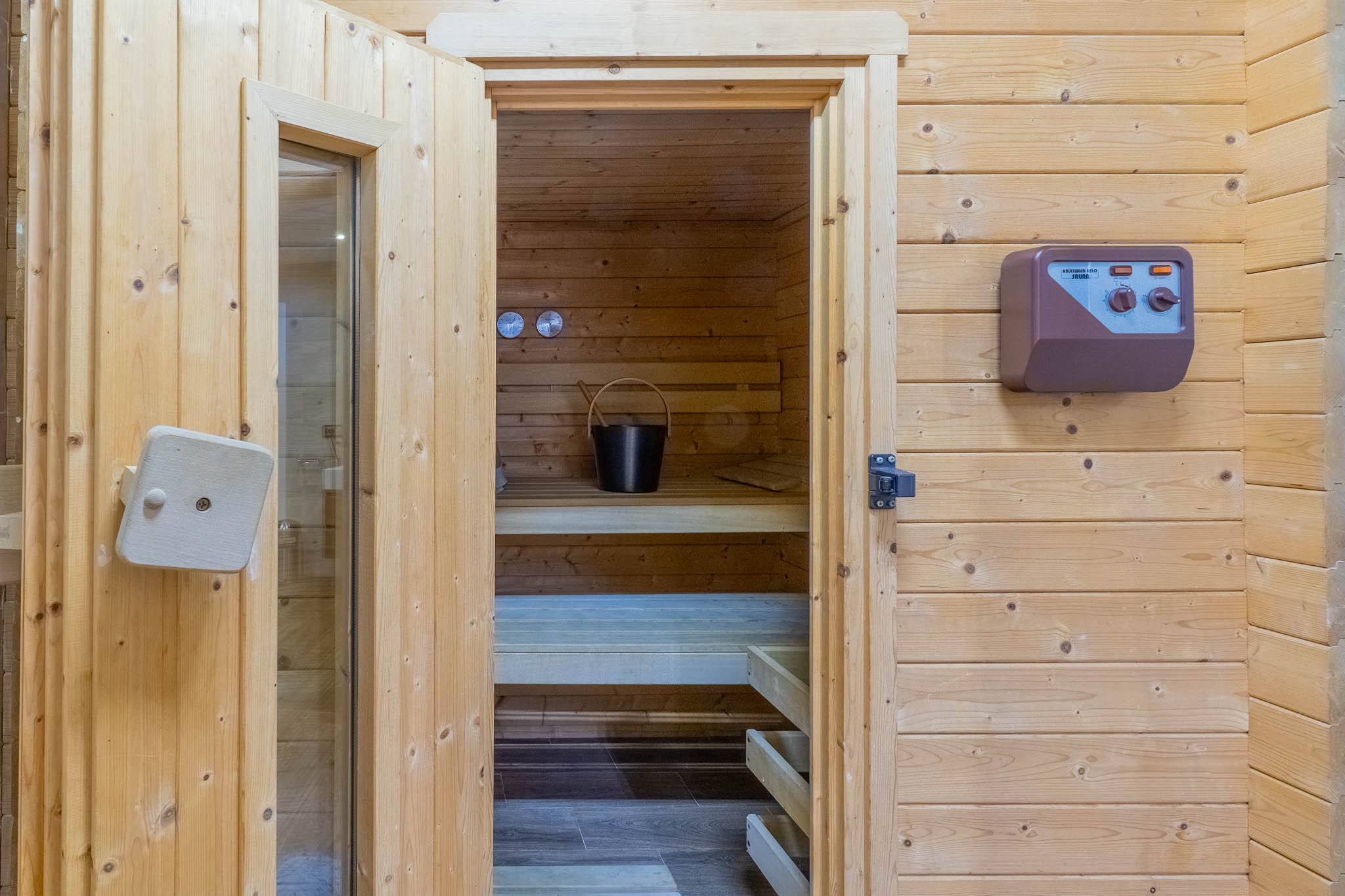 Accès au sauna en bois clair, avec ses bancs intérieurs, un seau et une louche, ainsi qu'un panneau de contrôle mural externe.