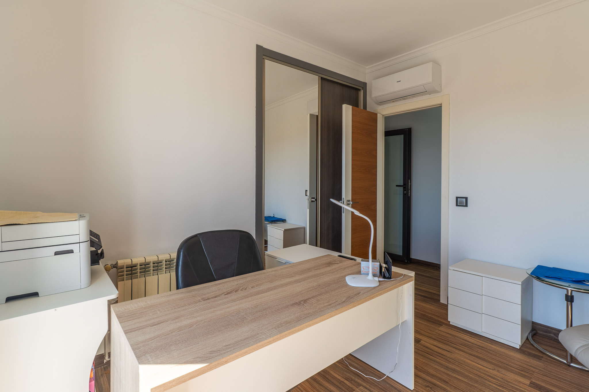 Bureau lumineux avec un bureau imitation bois clair, sol stratifié, armoire coulissante miroir et unité de climatisation murale.