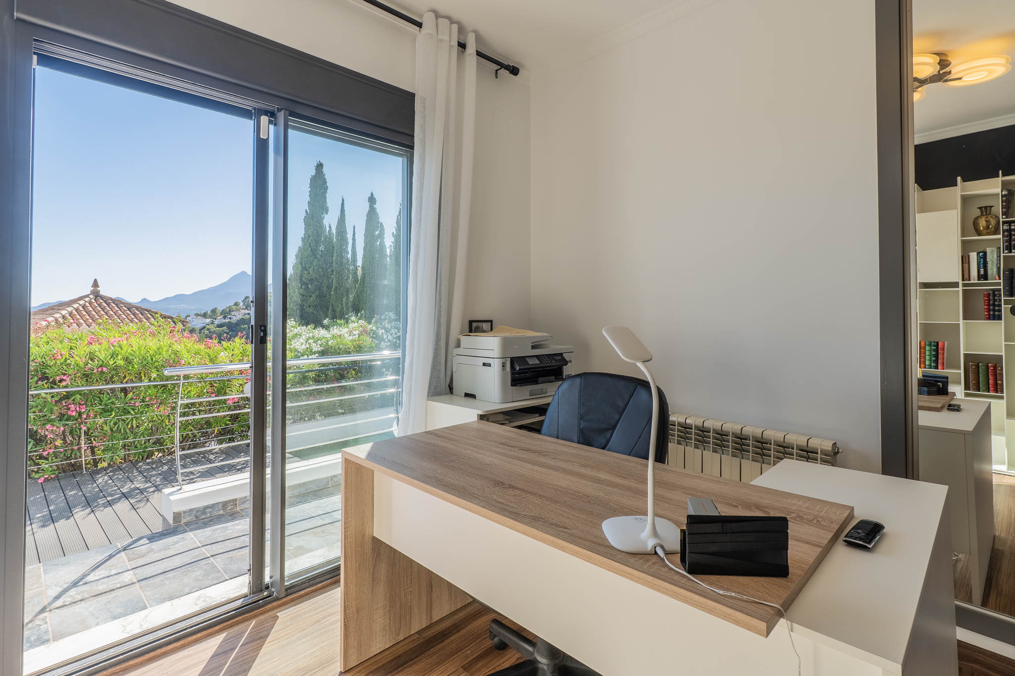 Bureau lumineux avec un bureau moderne en bois, une grande porte coulissante vitrée s'ouvrant sur une terrasse verdoyante et des vues panoramiques sur la montagne. Un miroir reflète une étagère fonctionnelle.