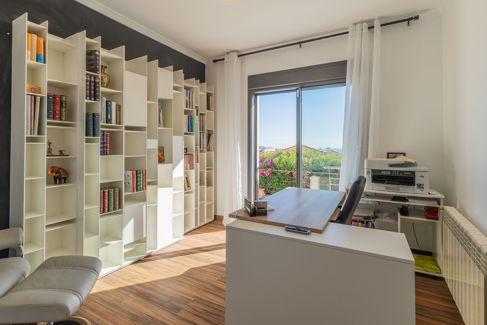 Bureau lumineux avec une bibliothèque modulaire blanche, un sol en bois et un accès à un balcon offrant des vues sur la verdure. Le bureau moderne est baigné de lumière naturelle.