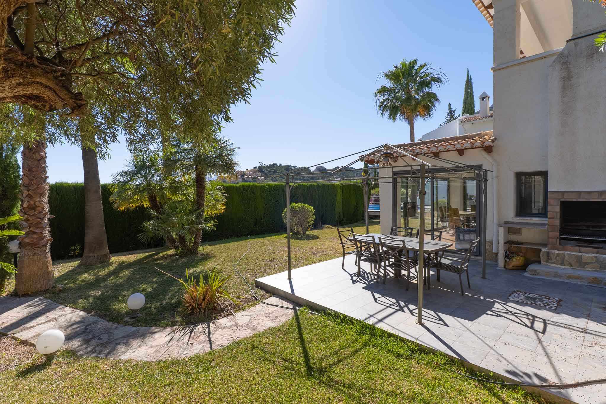 Cet espace repas extérieur comprend un patio carrelé sous une pergola métallique, parfait pour les repas en plein air. Il est complété par un barbecue intégré et un jardin luxuriant avec palmiers et haie.
