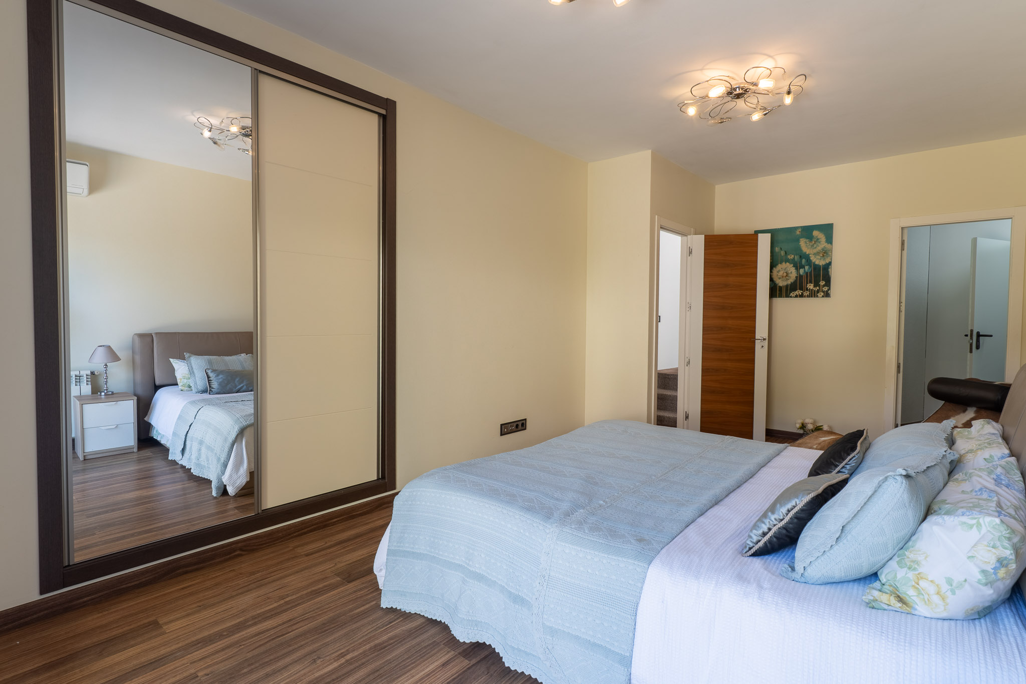 Chambre à coucher avec lit aux textiles bleus clairs, parquet stratifié et armoire coulissante à panneau miroir. Un luminaire de plafond moderne et un tableau floral complètent l'espace.