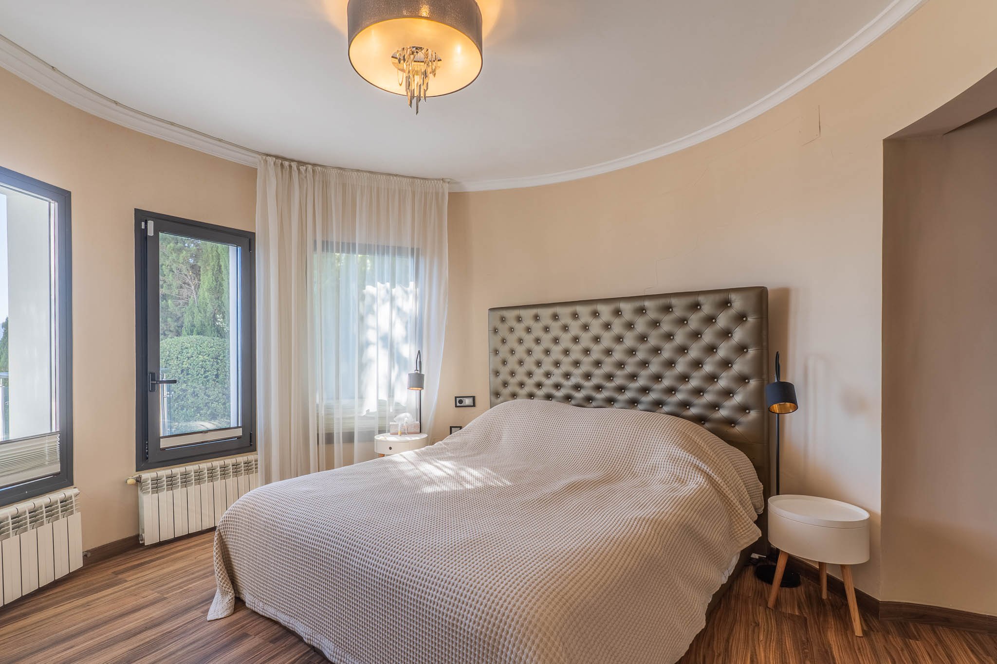 Chambre avec grand lit et tête de lit capitonnée. Mur incurvé avec fenêtres à cadre sombre et voilages clairs. Parquet, radiateurs et luminaire de plafond moderne.
