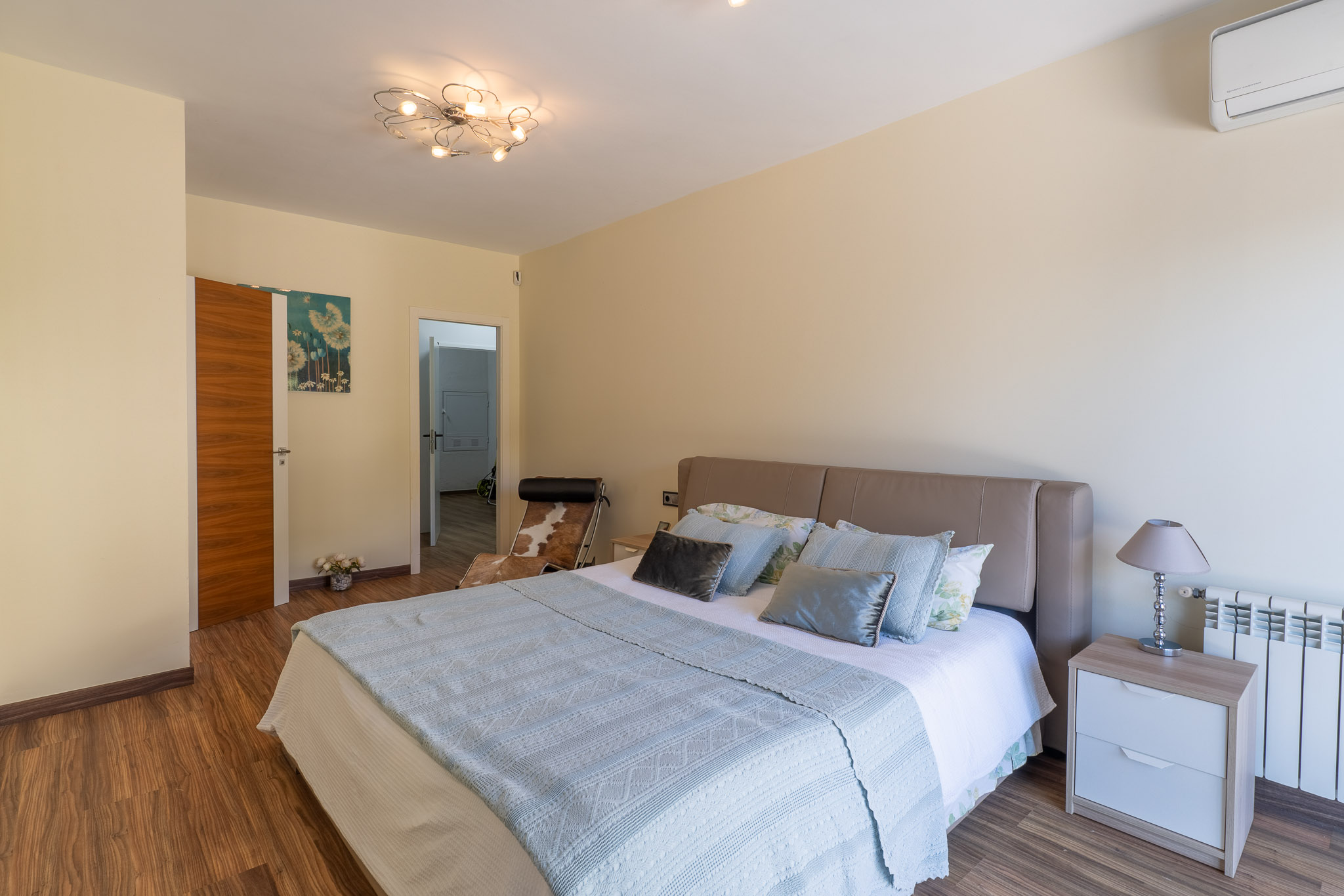 Chambre avec lit à tête de lit beige, parquet stratifié et luminaire de plafond moderne. Une table de chevet avec lampe, un radiateur et la climatisation sont visibles.