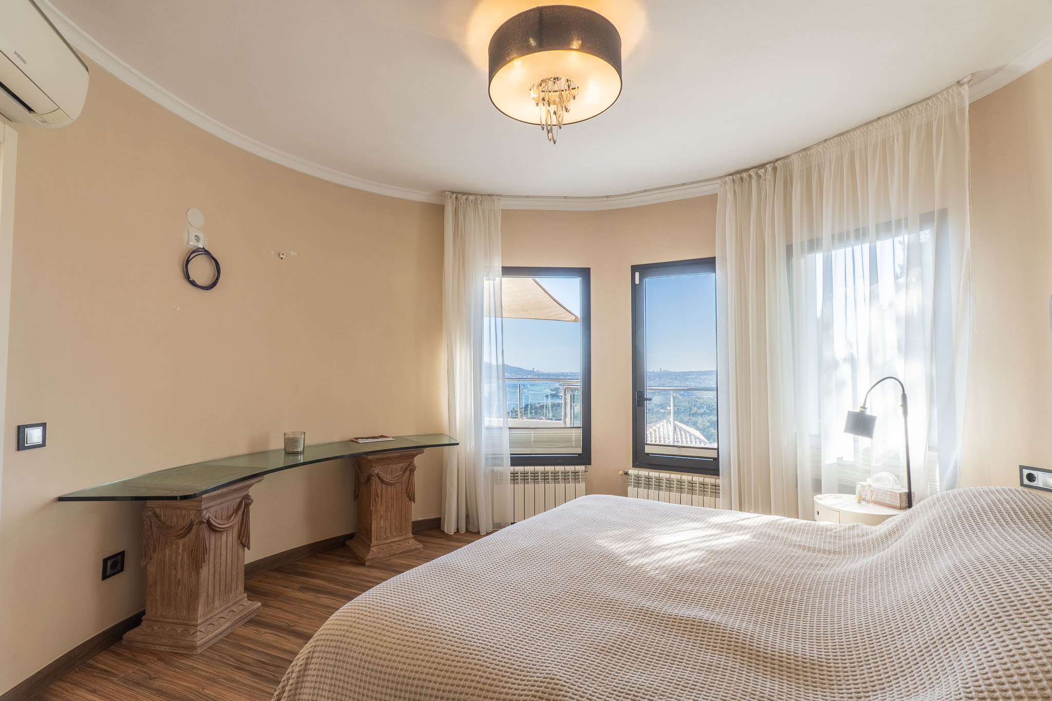 Chambre lumineuse avec vues panoramiques sur la mer, grandes fenêtres à voilages et un bureau en verre sur des socles en bois sculpté.