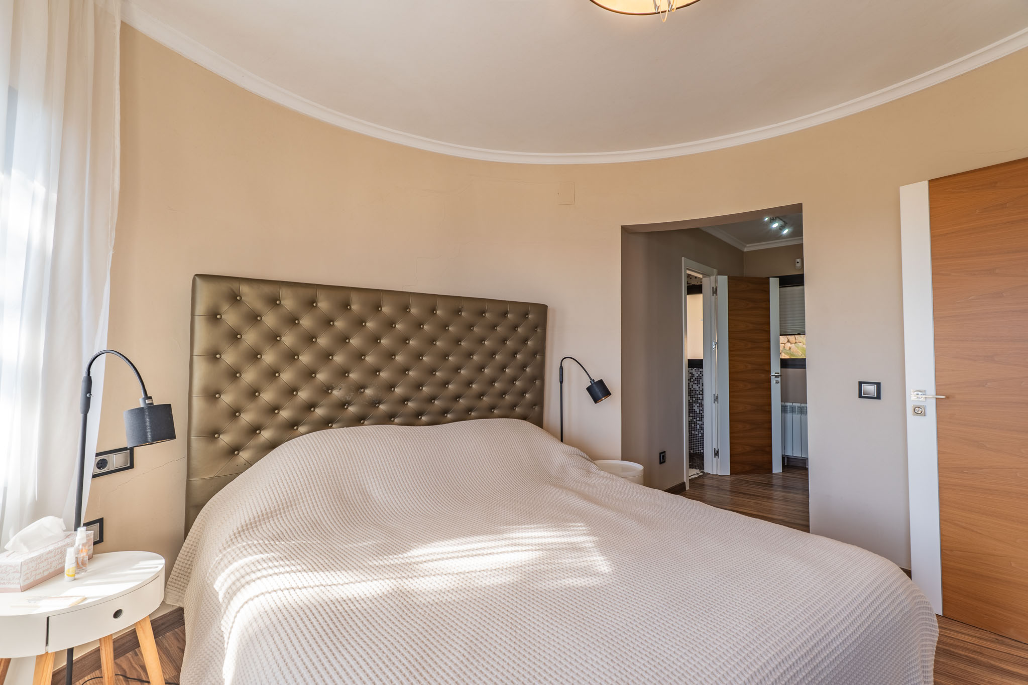 Chambre principale avec tête de lit capitonnée, mur incurvé et lumière naturelle filtrée par des voilages, complétée par des portes en bois.