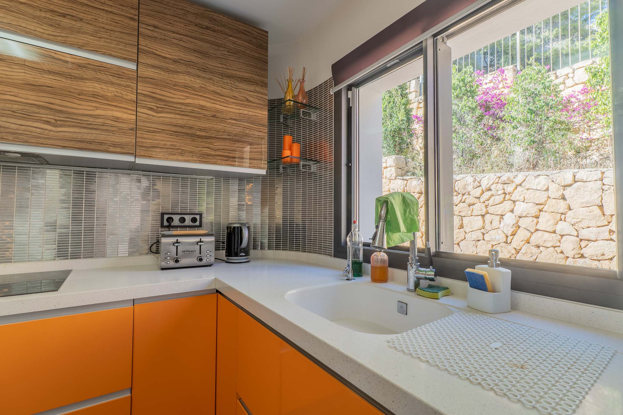 Cuisine moderne avec comptoir en quartz blanc, armoires basses orange vif et unités supérieures en bois veiné. Fenêtre offrant une vue sur un mur de pierre et la végétation.