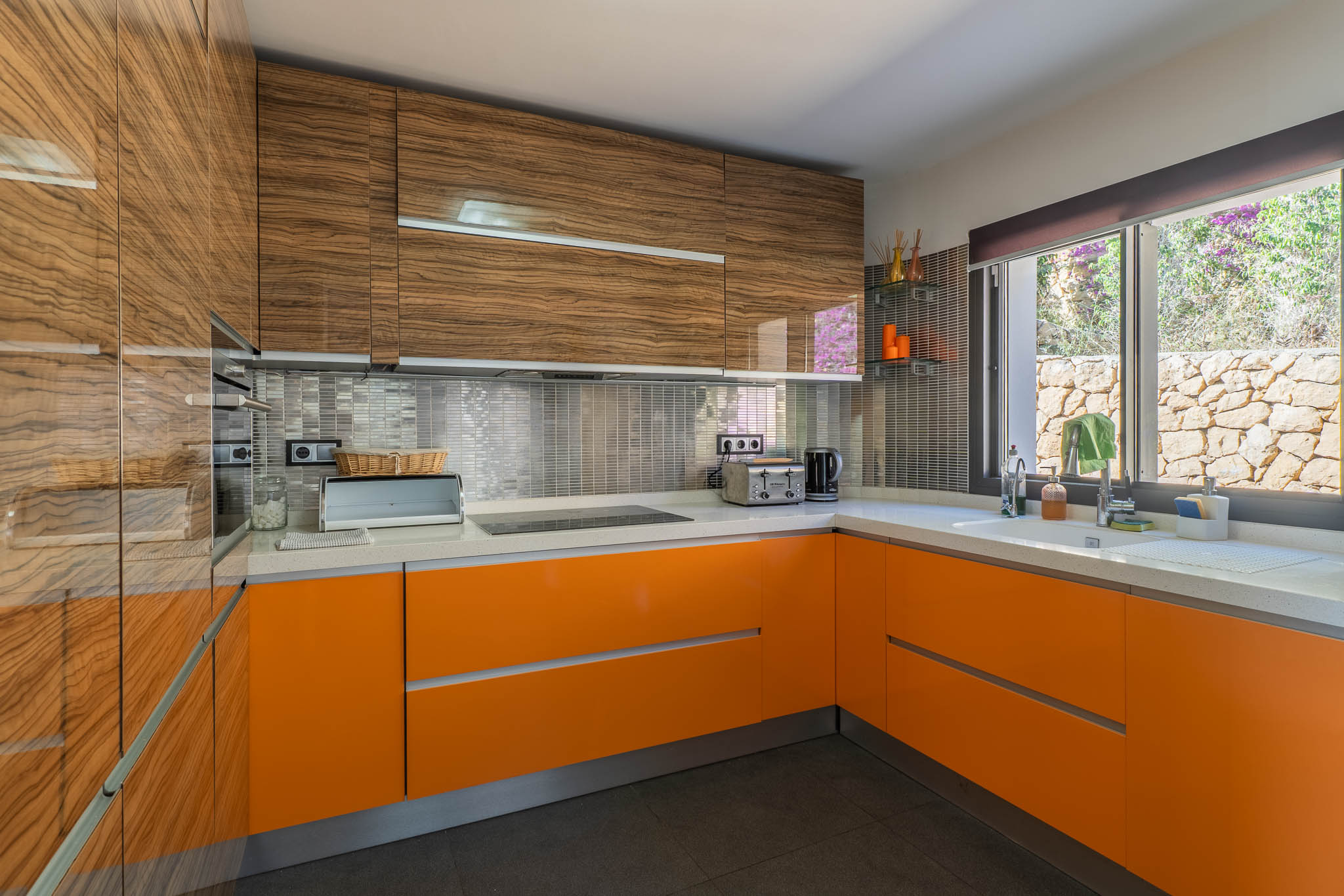 Cuisine moderne avec meubles hauts en bois veiné brillant et bas laqués orange vif. Le plan de travail blanc, la crédence en mosaïque métallique et la fenêtre donnant sur un mur de pierre sont visibles.