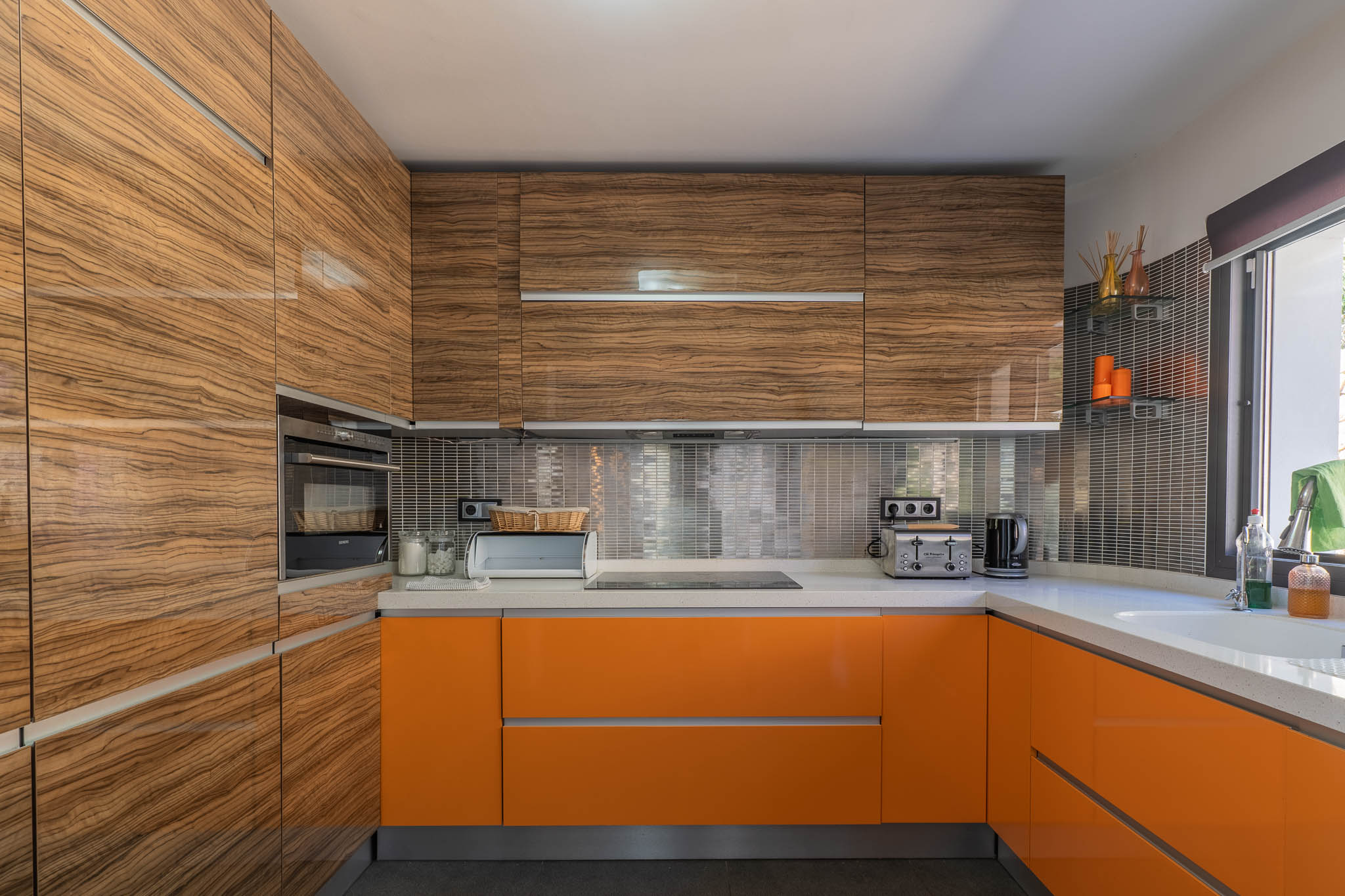 Cuisine moderne avec meubles hauts en bois veiné brillant et bas orange vif, complétée par des plans de travail clairs et une crédence en mosaïque métallique.