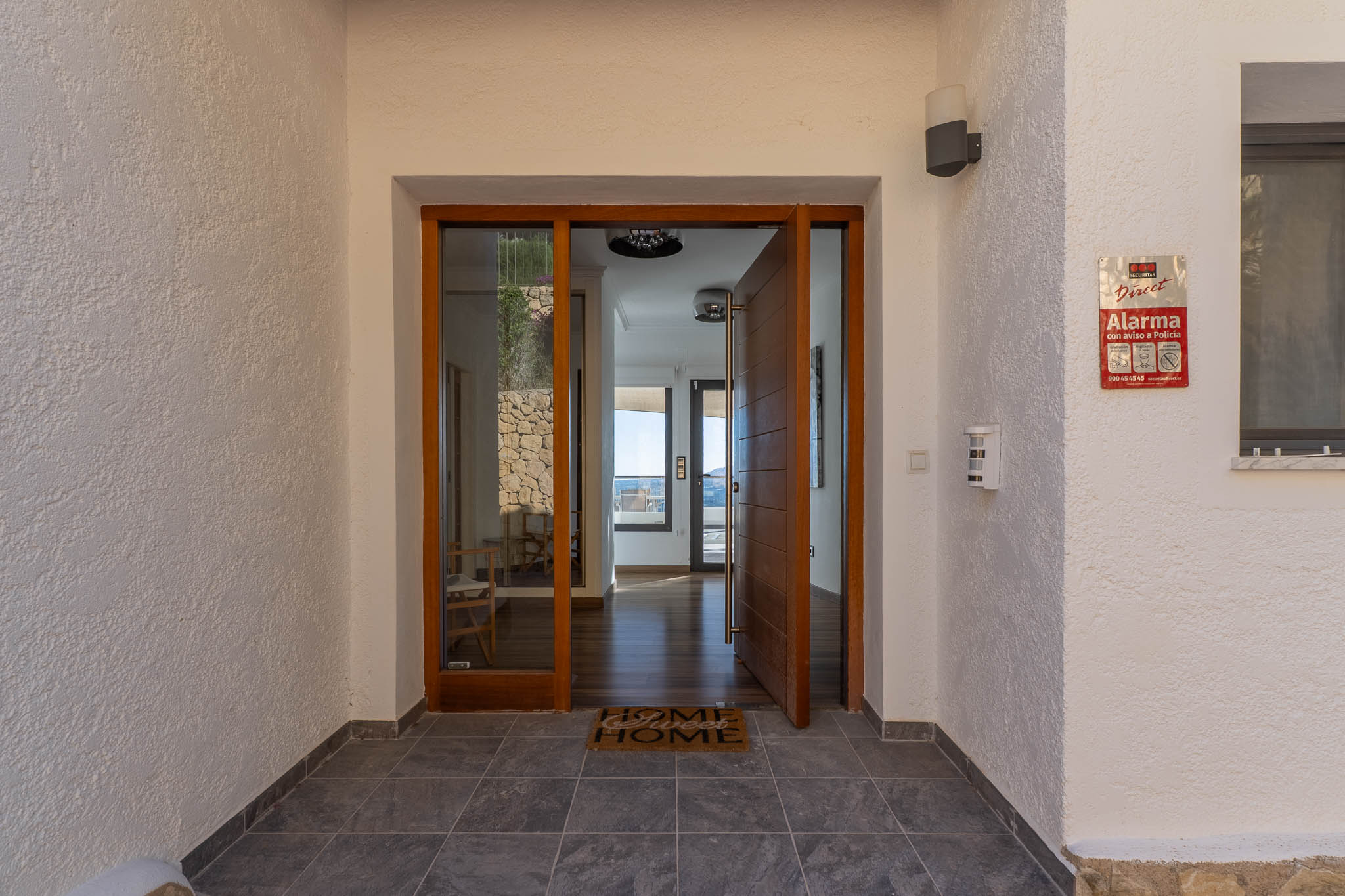 Entrée principale avec porte pivotante en bois et panneau vitré, ouvrant sur un intérieur lumineux aux parquets et vues mer. Murs extérieurs texturés et carrelage foncé.