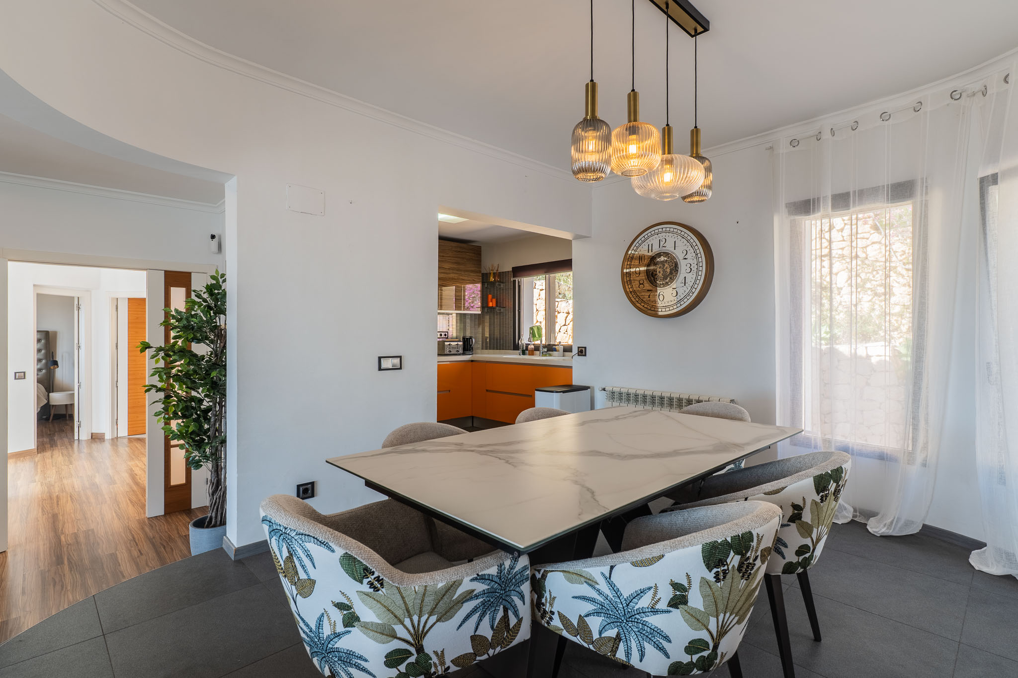 Espace repas avec table effet marbre et chaises à motif tropical. L'éclairage suspendu moderne complète la pièce, ouverte sur une cuisine aux armoires oranges.
