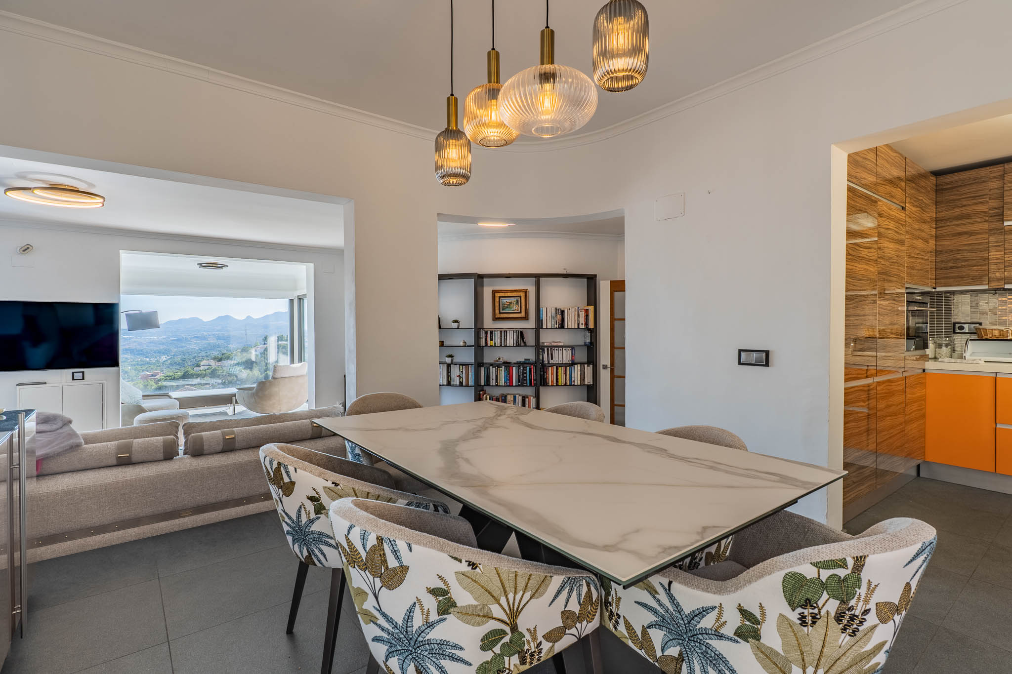 Espace salle à manger design avec table effet marbre et chaises tapissées, éclairé par des suspensions modernes. S'ouvre sur le salon et des vues panoramiques sur la montagne.