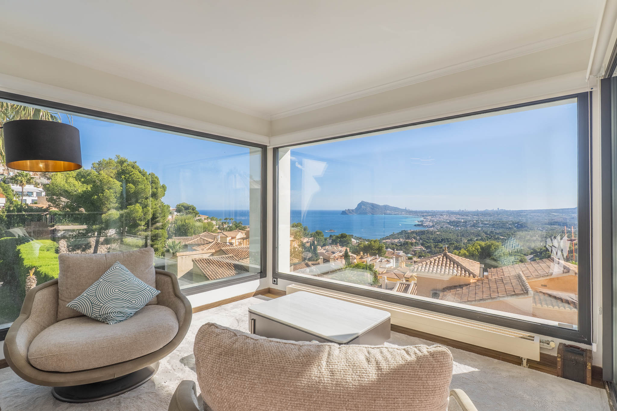 Espace salon avec de larges baies vitrées d'angle offrant une vue panoramique sur la mer et la côte. Deux fauteuils et une table basse moderne créent un lieu lumineux.