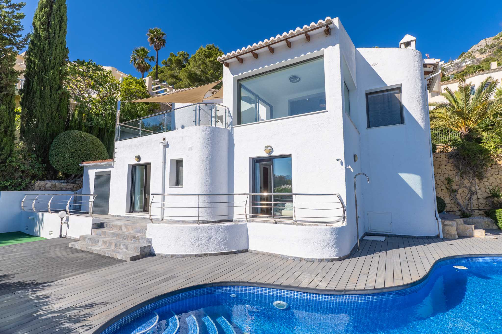 Extérieur de la villa blanche avec grandes baies vitrées et terrasses à balustrades en verre. La piscine en mosaïque bleue et le pont en composite gris sont entourés de cyprès et de palmiers.