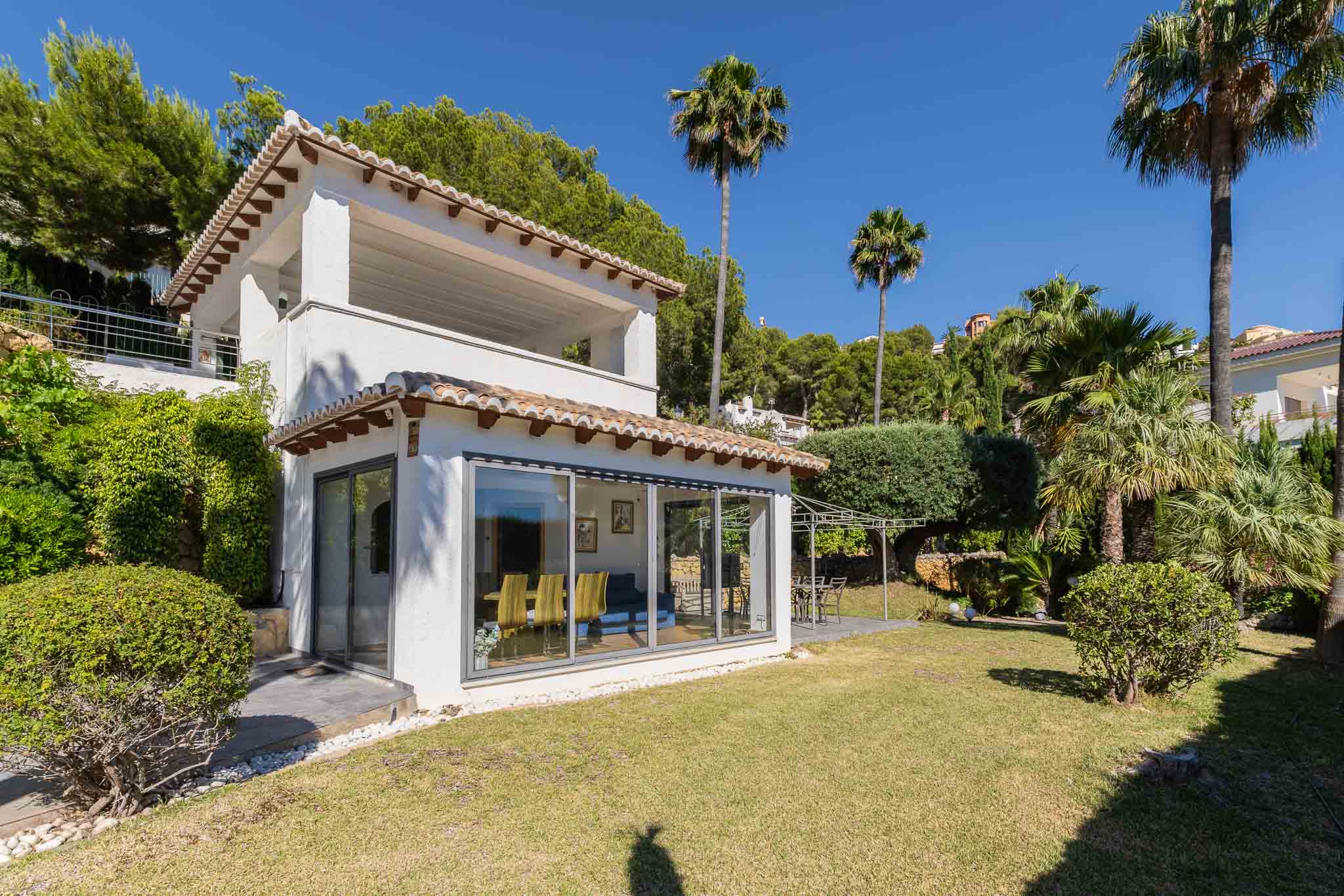 Façade blanche de la villa avec tuiles traditionnelles et grandes baies vitrées. Le jardin plat comprend une pelouse verdoyante et un patio avec pergola, entouré de palmiers et arbustes.