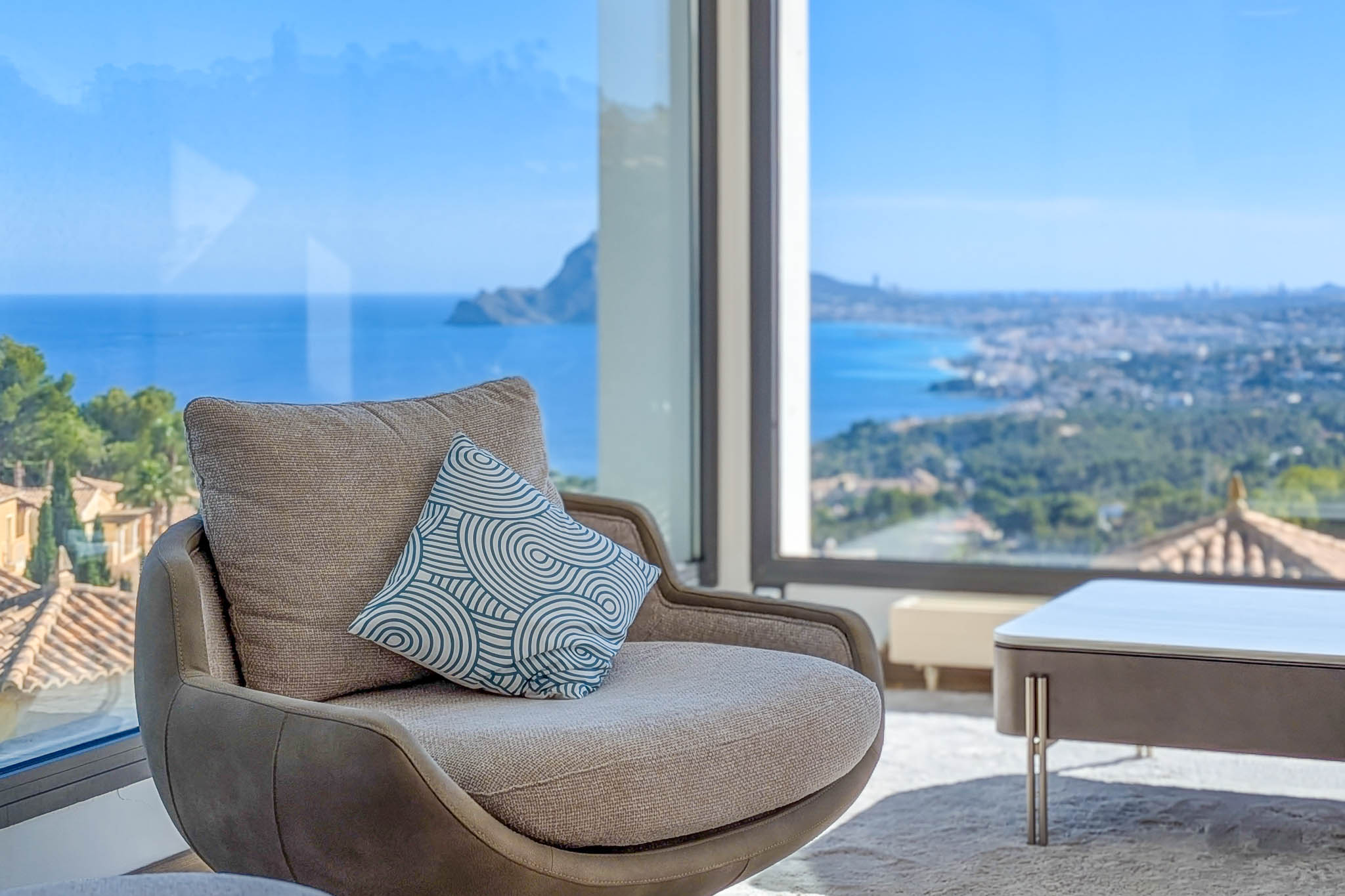 Fauteuil confortable devant une fenêtre panoramique, offrant des vues imprenables sur la mer Méditerranée et le littoral. Le coussin décoratif modernise l'espace intérieur.