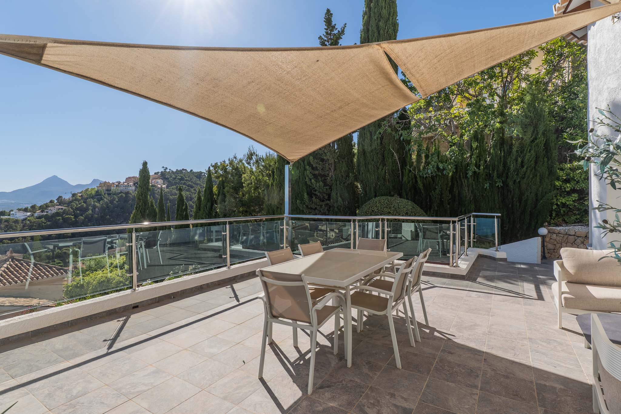 Grande terrasse extérieure ombragée par une voile solaire, meublée d'une table à manger et bordée d'une balustrade en verre, avec vue panoramique sur les montagnes.
