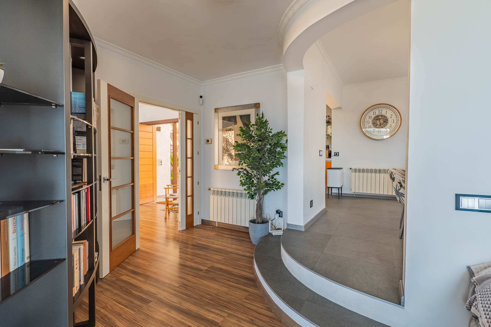 Hall d'entrée avec parquet stratifié effet bois, s'ouvrant sur un espace extérieur par des portes vitrées. Des marches carrelées courbes mènent à la salle à manger, avec une bibliothèque intégrée.