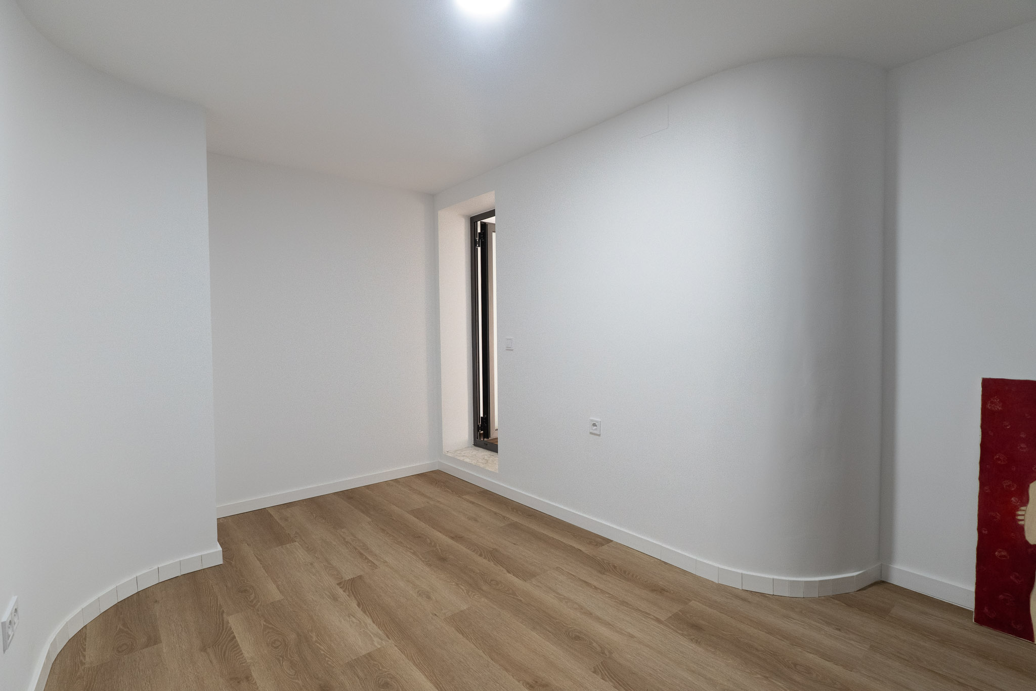 Pièce lumineuse avec sol en lames claires, murs blancs et une paroi courbe distinctive. Présente une ouverture verticale à cadre sombre et seuil en pierre.