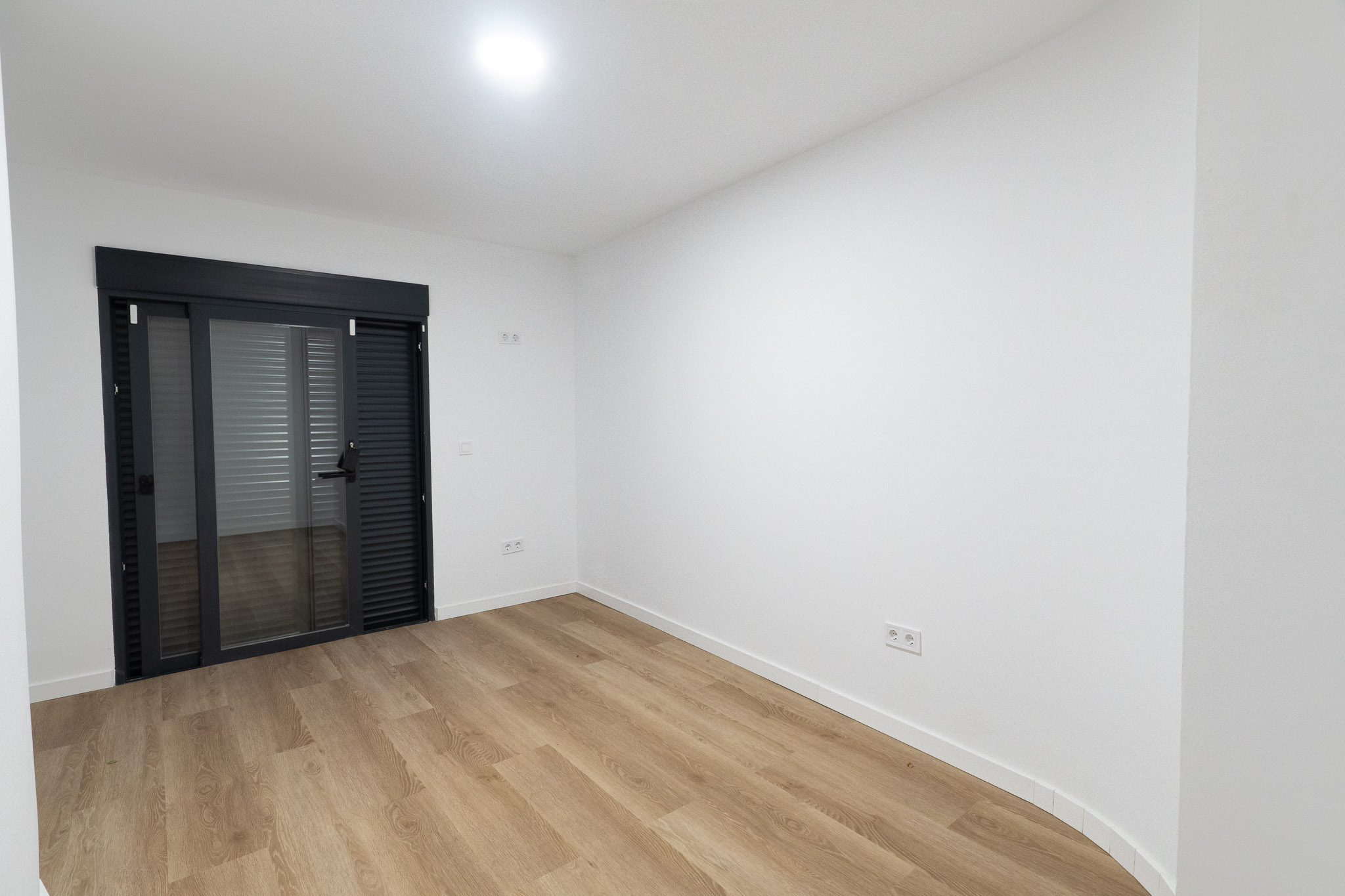 Pièce lumineuse avec un sol stratifié imitation bois clair et des murs blancs. Une grande porte coulissante en verre gris anthracite avec stores intégrés mène à l'extérieur.