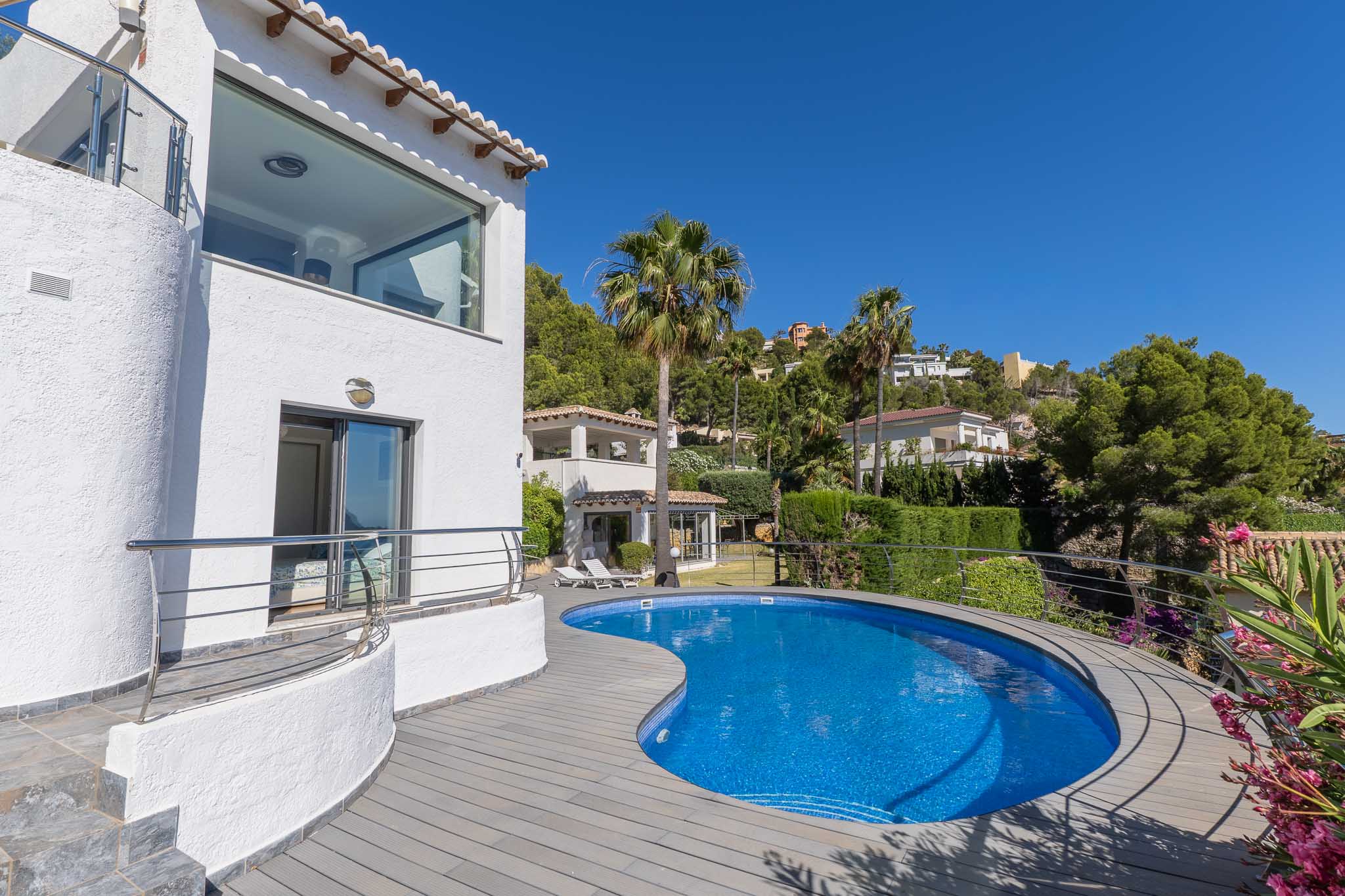 Piscine courbe en mosaïque bleue avec terrasse en composite gris, jouxtant une façade blanche dotée d'une fenêtre panoramique et d'un balcon à balustrade en acier.