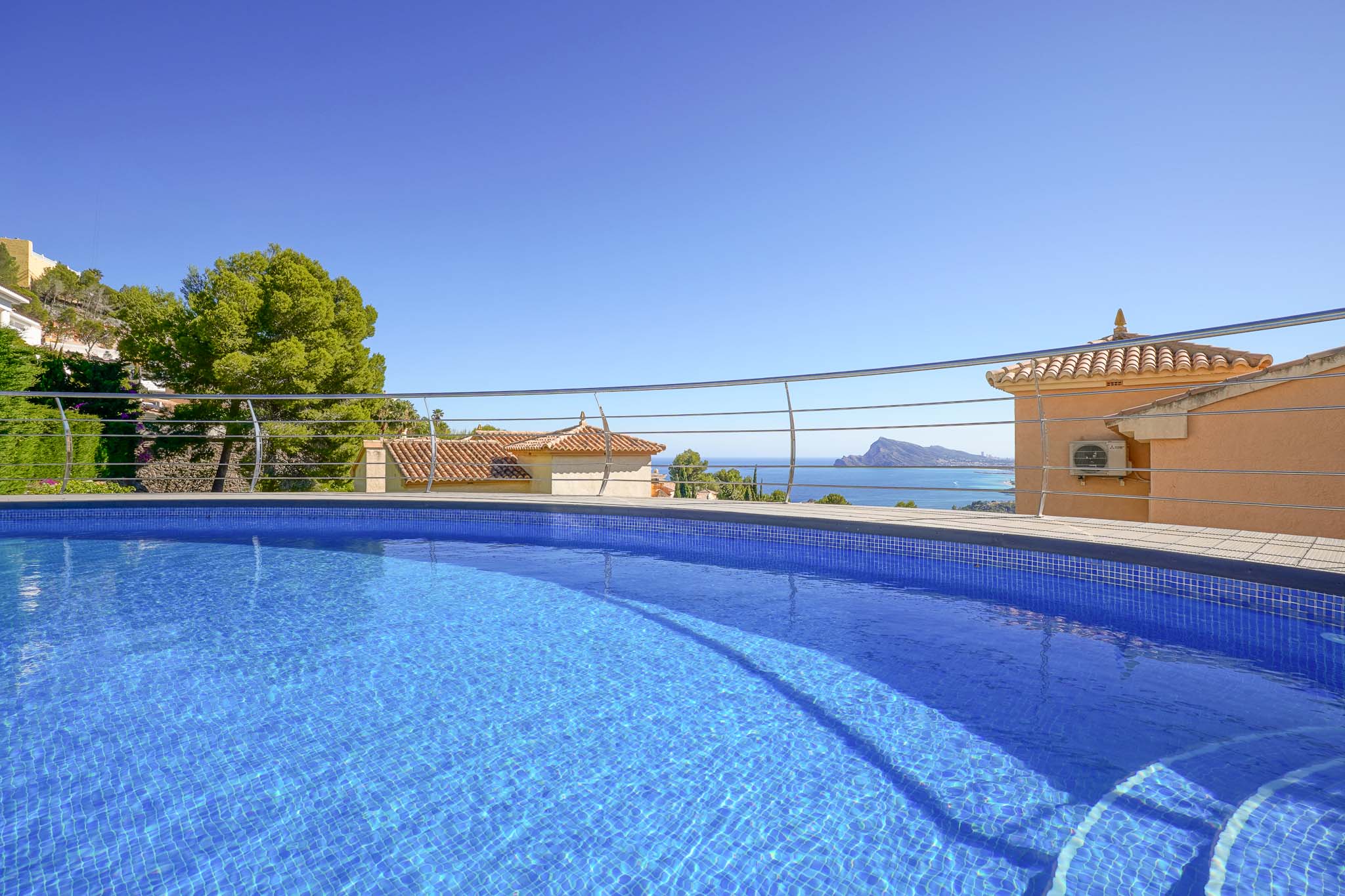 Piscine en mosaïque bleue avec escaliers intégrés et balustrade moderne, encadrant des vues panoramiques sur la mer, des toits traditionnels et la côte.