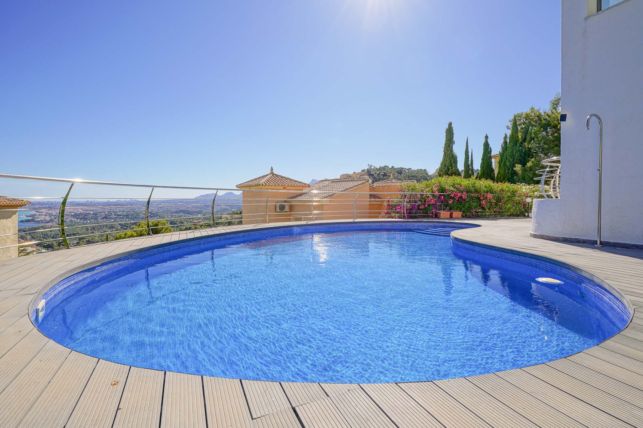Piscine incurvée en mosaïque bleue, entourée d'une terrasse en bois clair, avec une balustrade en acier inoxydable et vue panoramique sur la vallée et la ville.