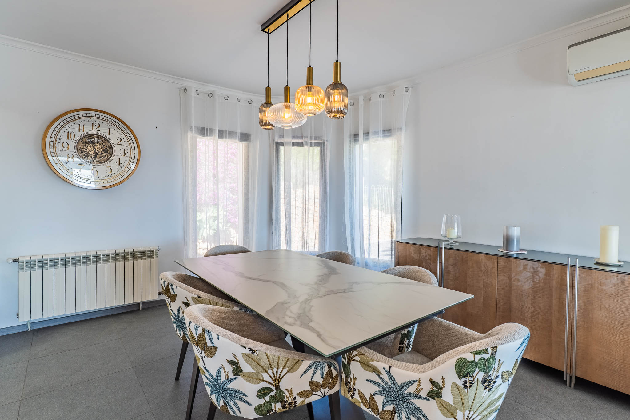 Salle à manger lumineuse avec table effet marbre, chaises à motif tropical et lustre linéaire moderne, baignée de lumière naturelle.