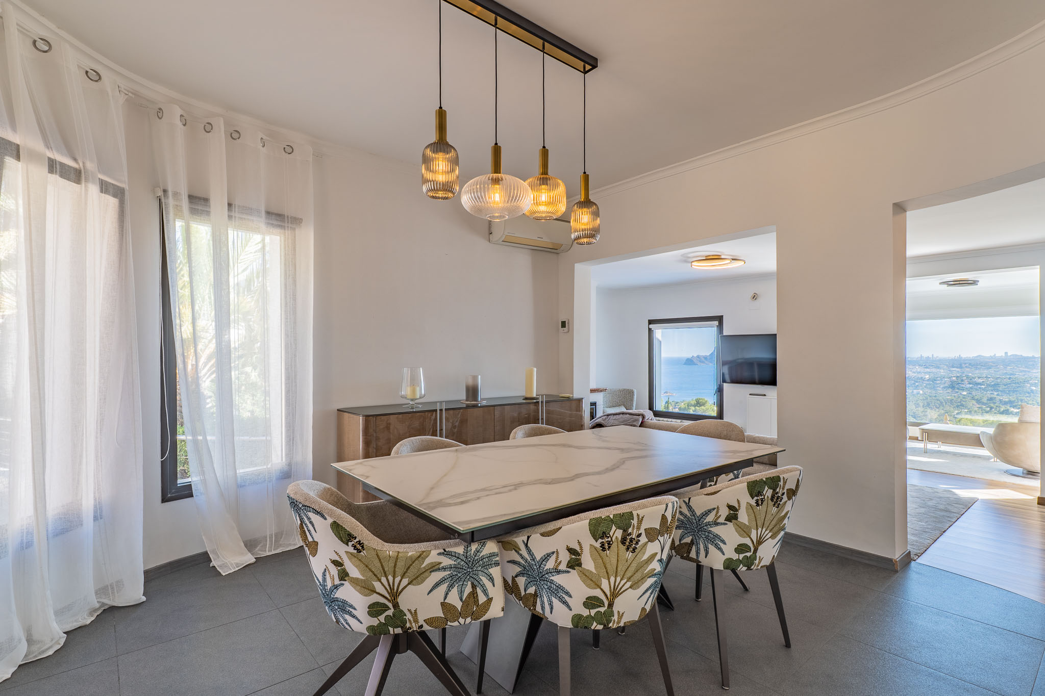 Salle à manger moderne avec table effet marbre et chaises à motif tropical. Un lustre linéaire éclaire l'espace, qui s'ouvre sur des vues panoramiques sur la mer depuis les pièces adjacentes.