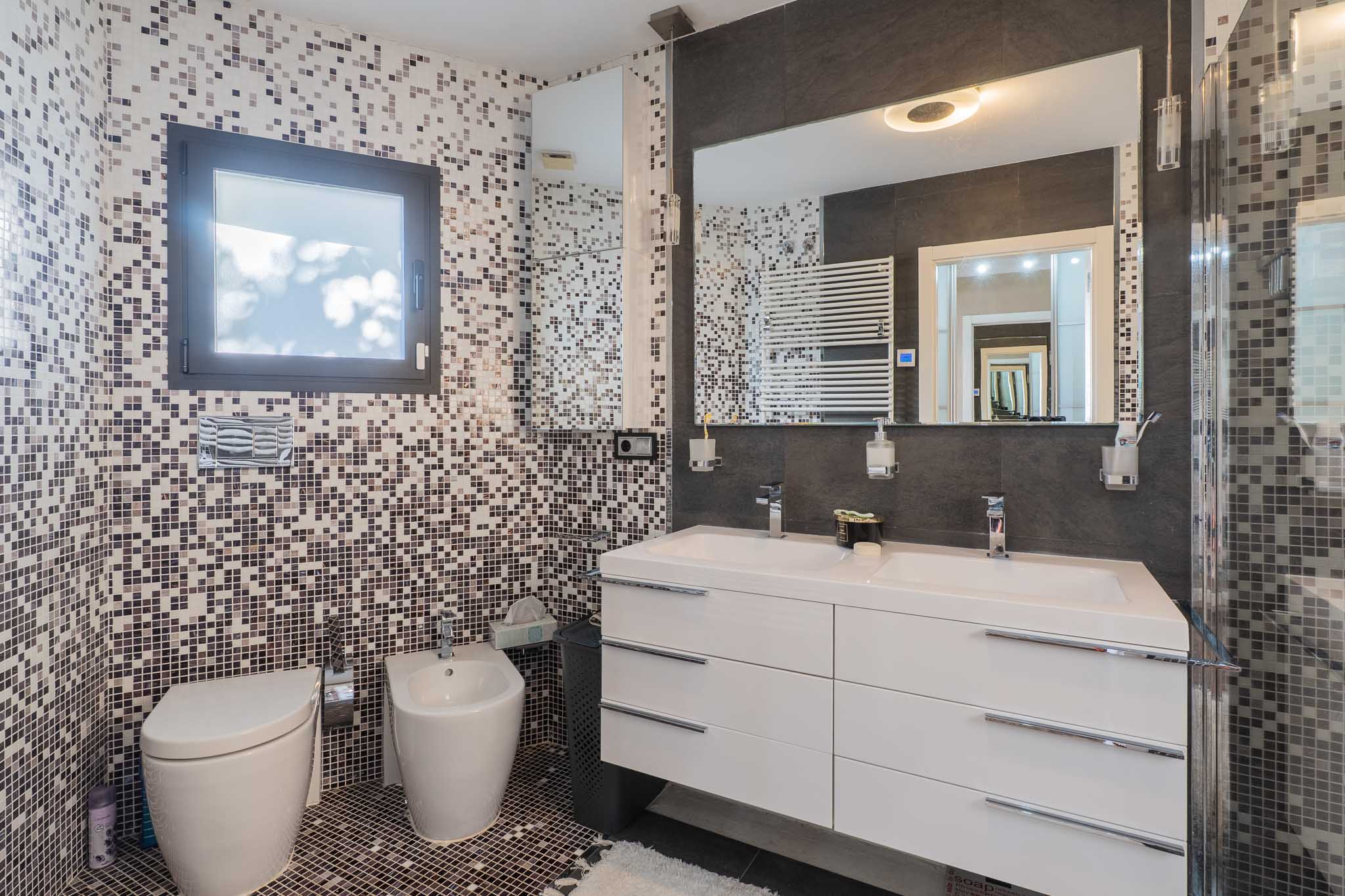 Salle de bain contemporaine avec murs et sol en mosaïque tons terre/gris. Double vasque blanche, grand miroir éclairé, WC et bidet suspendus, plus un sèche-serviettes mural.