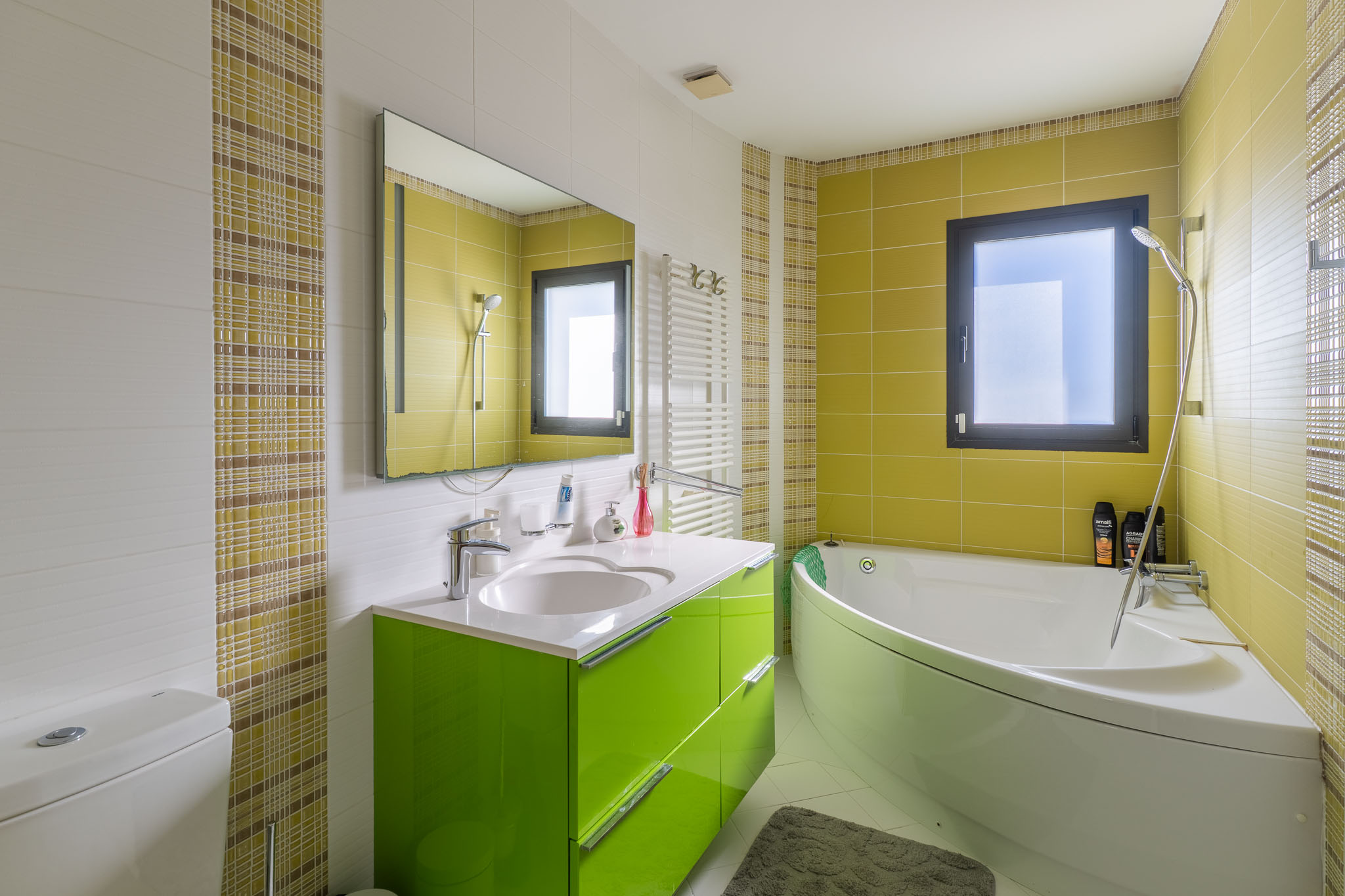Salle de bain moderne avec baignoire d'angle et meuble-lavabo vert vif. Carrelage mural blanc texturé, vert lime et mosaïque à carreaux. Fenêtre et sèche-serviettes mural.