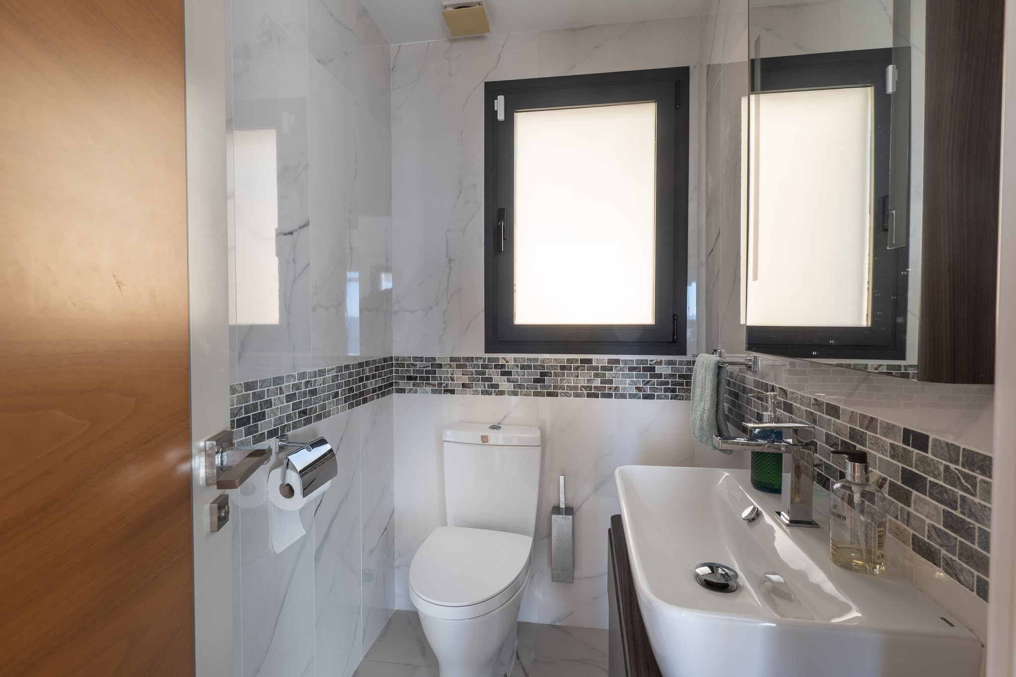 Salle de bain moderne avec carrelage effet marbre blanc, une frise mosaïque grise et une fenêtre à cadre sombre. Comprend un lavabo blanc et des toilettes assorties.