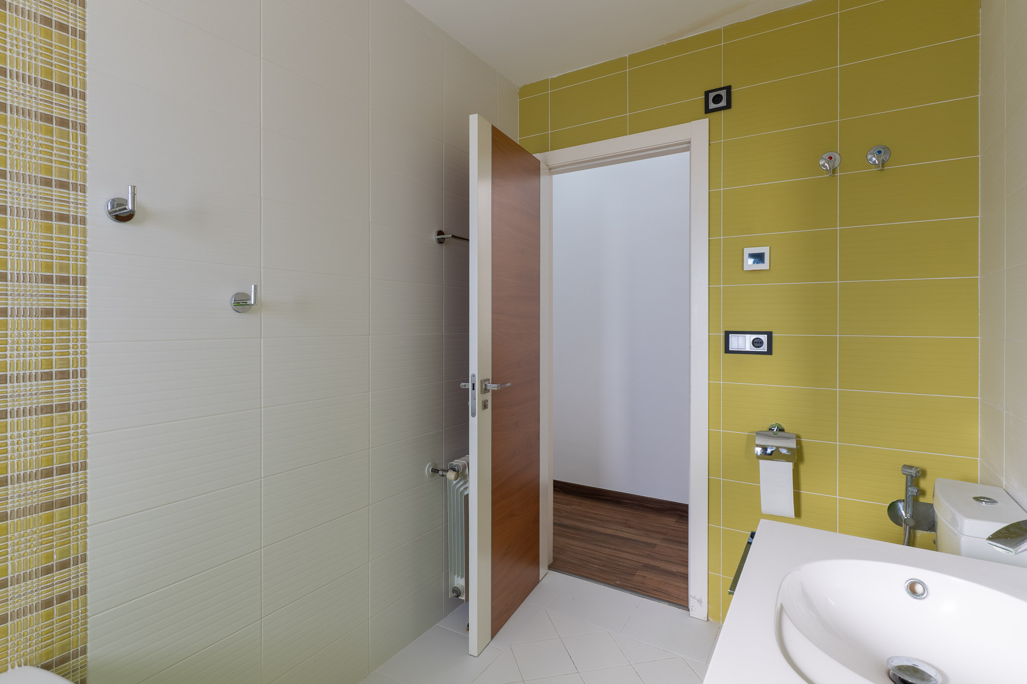 Salle de bain moderne avec carrelage mural vert anis et beige texturé, agrémenté d'une bande mosaïque. Elle est équipée d'un lavabo contemporain et d'une porte en bois.
