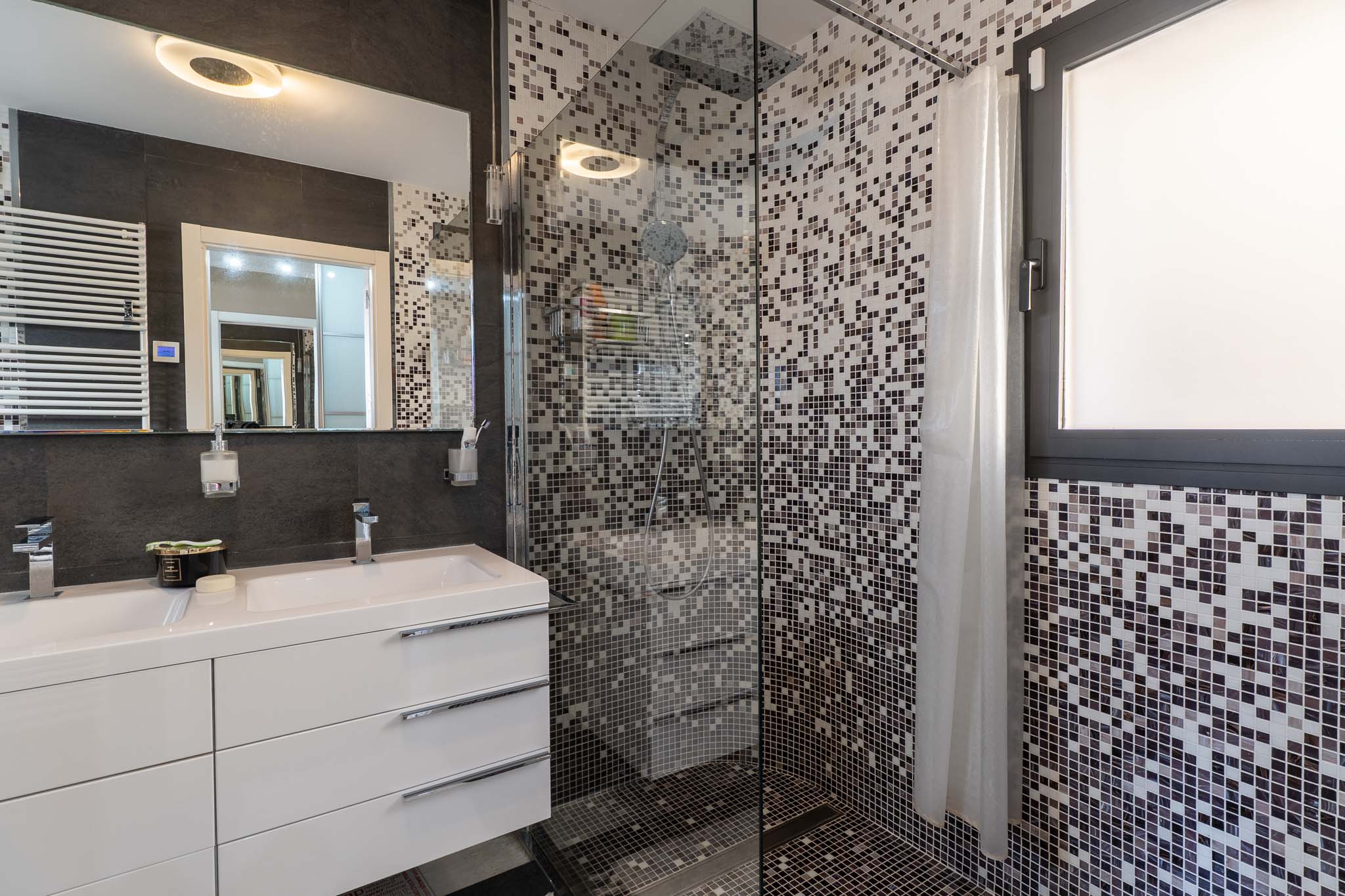 Salle de bain moderne avec double vasque, meuble blanc et miroir éclairé. La douche vitrée est revêtue de mosaïque grise et inclut une fenêtre intégrée.