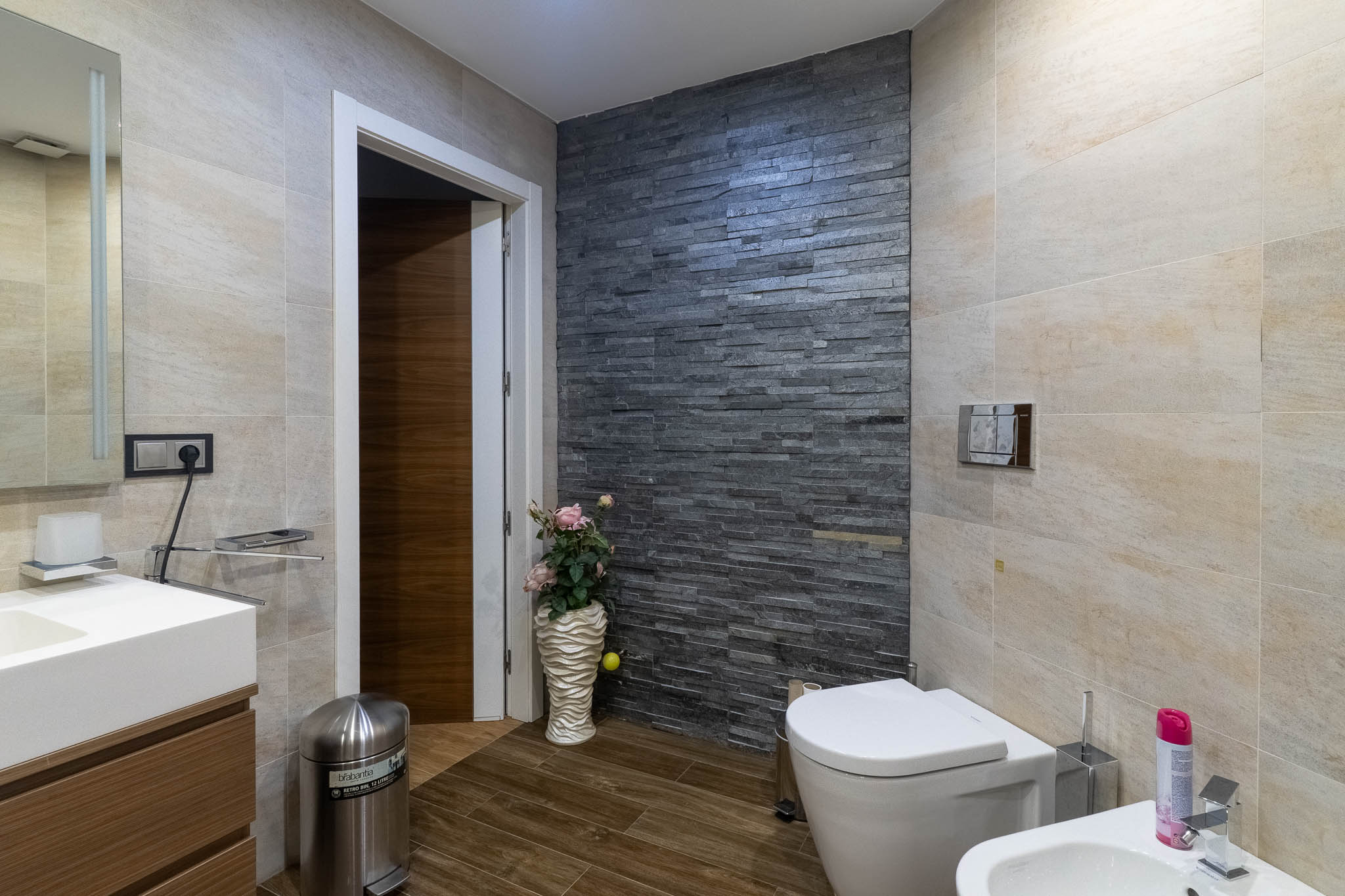 Salle de bain moderne avec mur d'accent en pierre empilée foncée, carrelage beige clair et sol imitation bois. Comprend toilettes, bidet et meuble-lavabo.