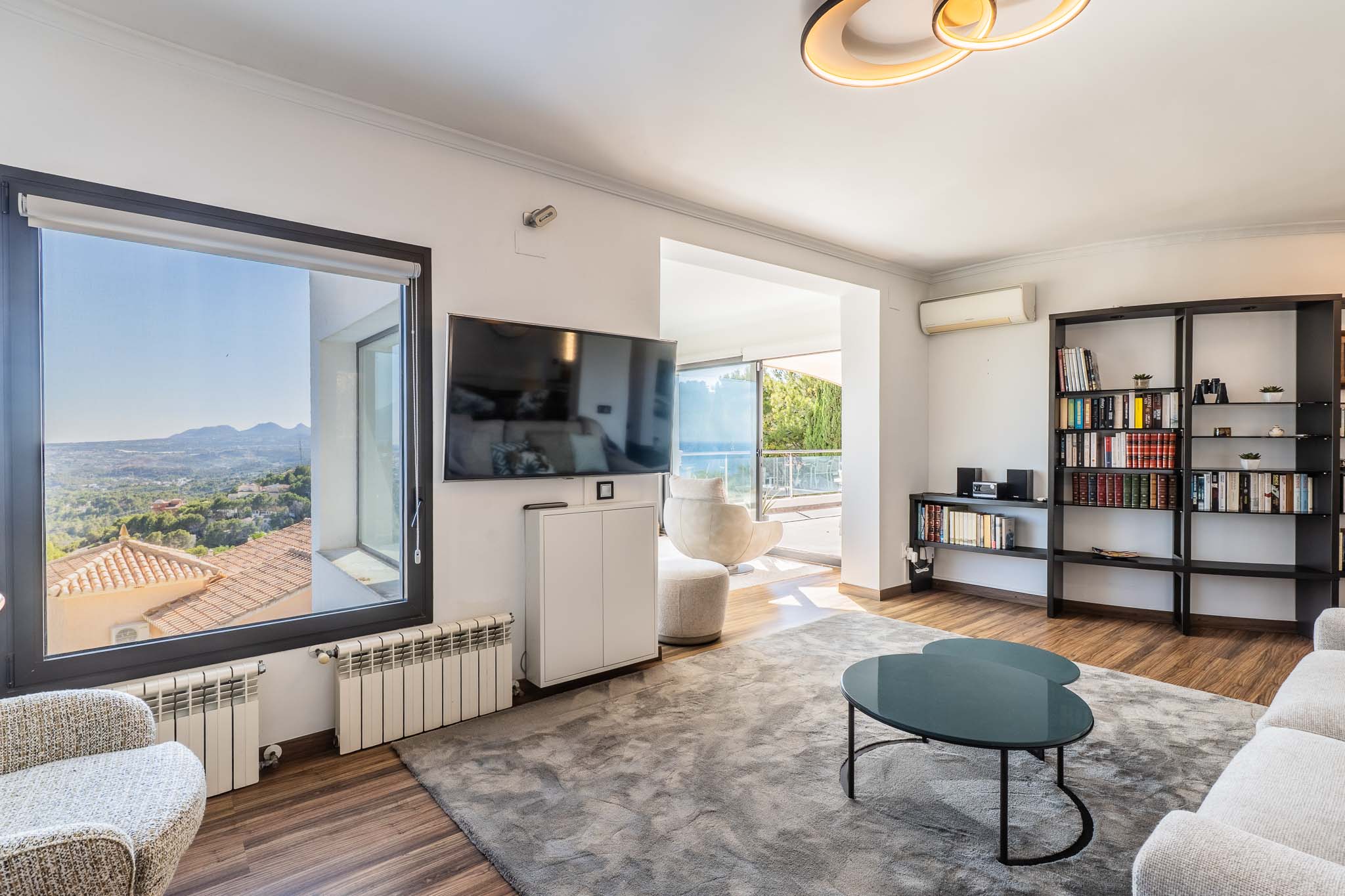 Salon lumineux offrant des vues panoramiques sur la montagne depuis la fenêtre et un accès à la terrasse avec vue sur mer. Il présente un sol en bois, un grand tapis gris et une bibliothèque moderne.
