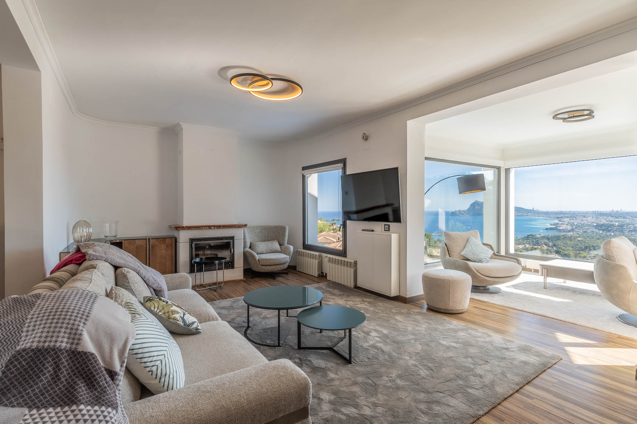 Salon moderne avec canapé clair, cheminée et larges baies vitrées offrant des vues panoramiques sur la mer et la ville. Le mobilier contemporain illumine l'espace.
