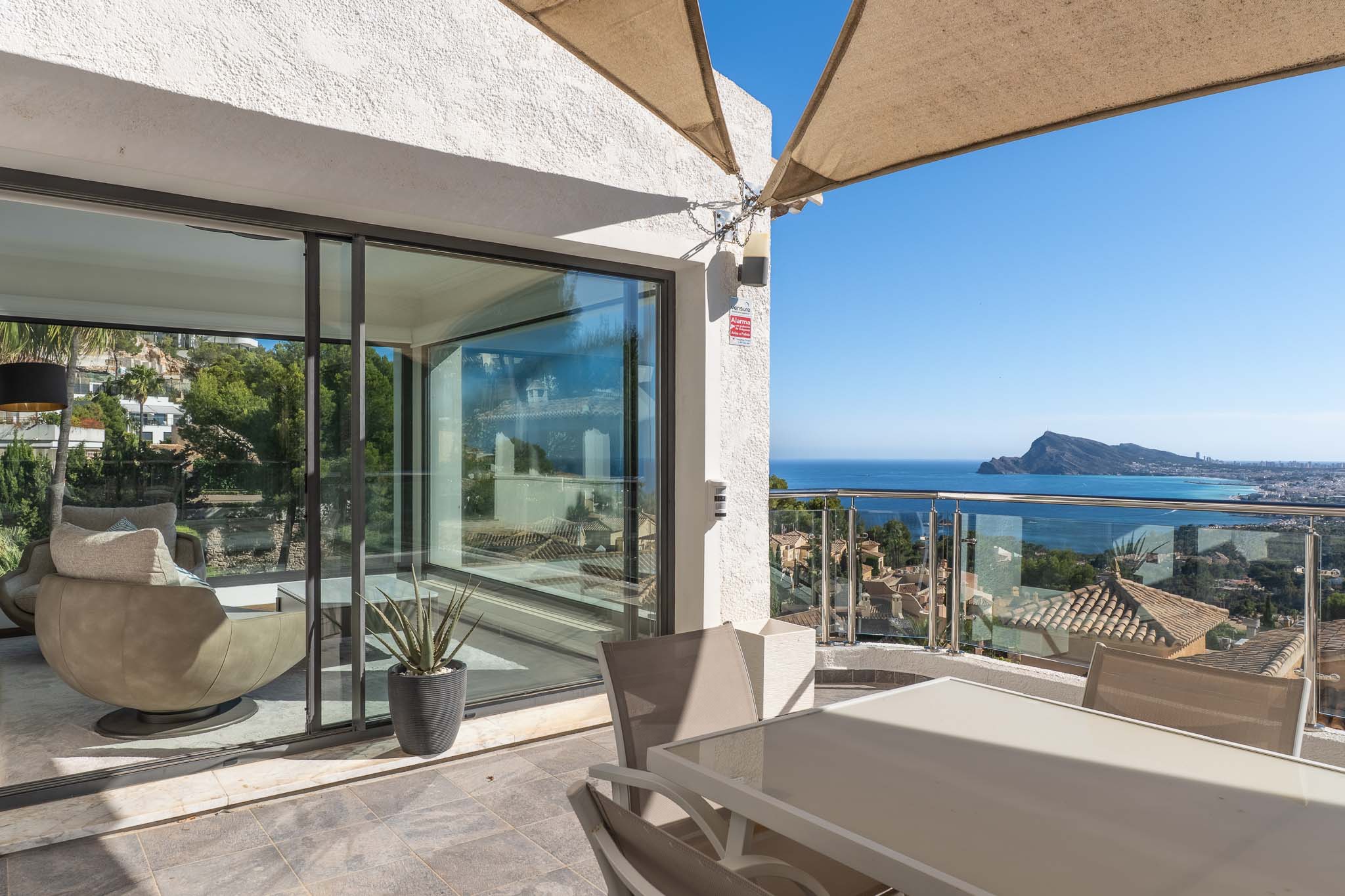 Terrasse extérieure aménagée avec table et chaises, sous une voile d'ombrage. Elle offre des vues panoramiques sur la mer et la côte via une balustrade en verre, ouvrant sur un intérieur lumineux.