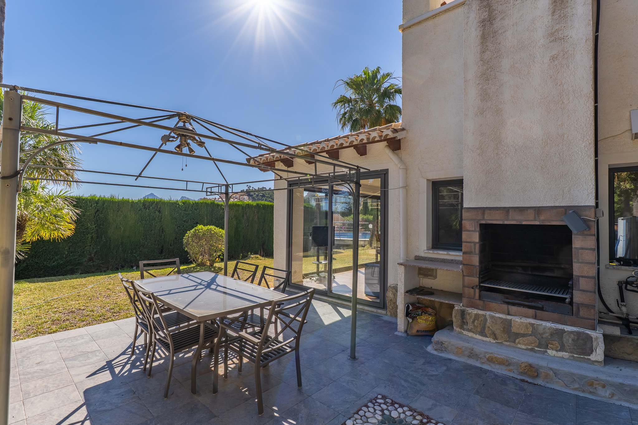 Terrasse extérieure carrelée en gris foncé, avec pergola métallique et table à manger. Un barbecue intégré en brique et pierre et des portes vitrées coulissantes sont visibles.