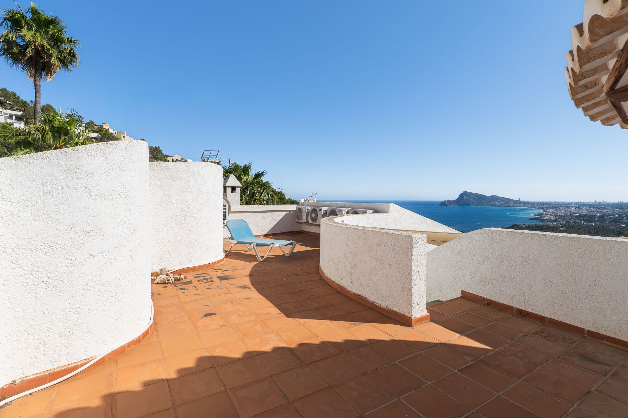 Terrasse sur le toit avec des carreaux de terre cuite et des murs blancs texturés, offrant une vue panoramique sur la mer et la côte. Une chaise longue bleue invite à la détente.
