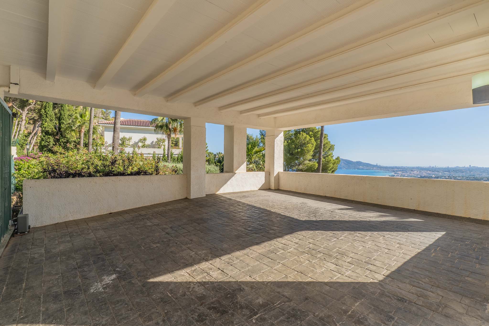 Vaste abri pour voitures couvert avec sol en béton estampé et plafond à poutres blanches. Il offre une vue panoramique sur la mer Méditerranée et la côte, encadrée de verdure luxuriante.