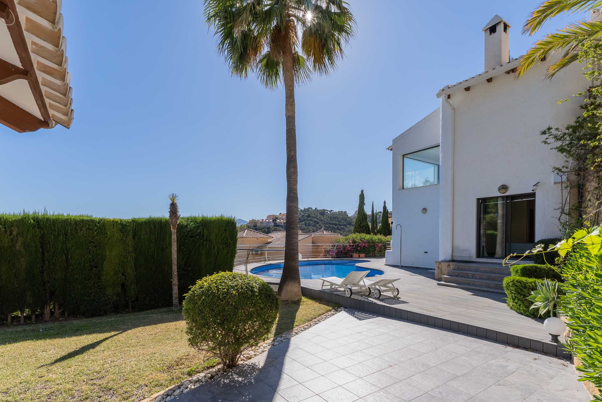 Vaste jardin avec piscine circulaire et solarium en bois, bordé par une villa blanche et une haie dense. Vues sur les collines environnantes.