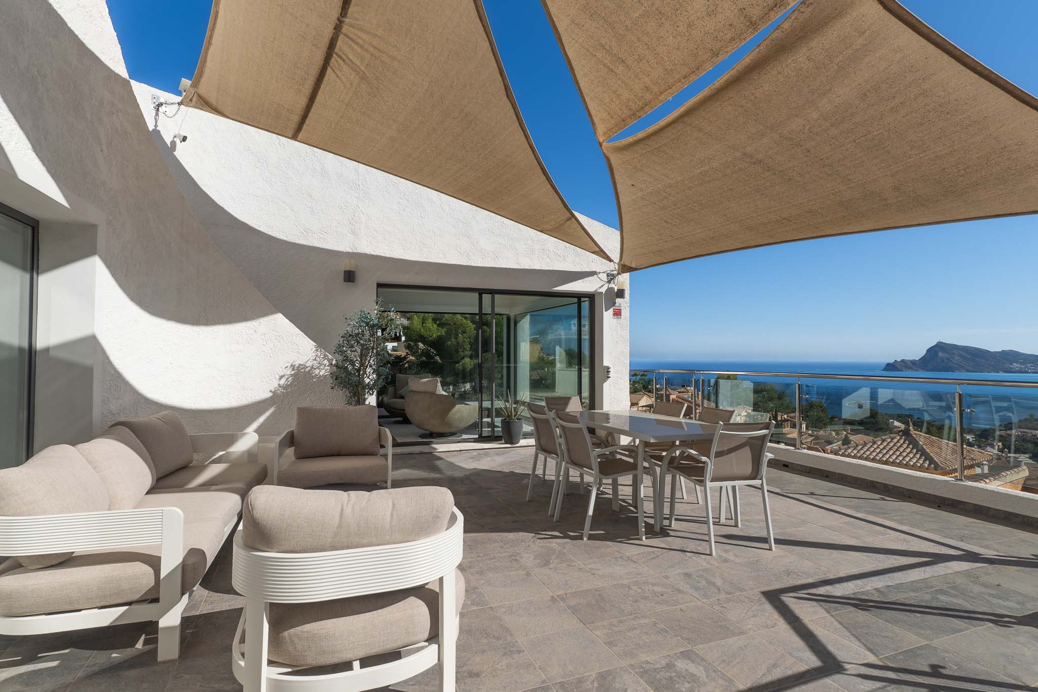 Vaste terrasse extérieure avec salon et salle à manger sous voile d'ombrage, offrant une vue panoramique sur la mer et la côte via une balustrade en verre.