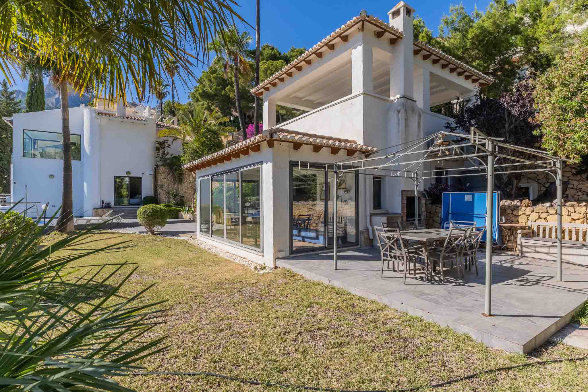Vaste terrasse pavée avec salle à manger extérieure sous pergola métallique, jouxtant une pièce vitrée et un jardin méditerranéen entretenu.